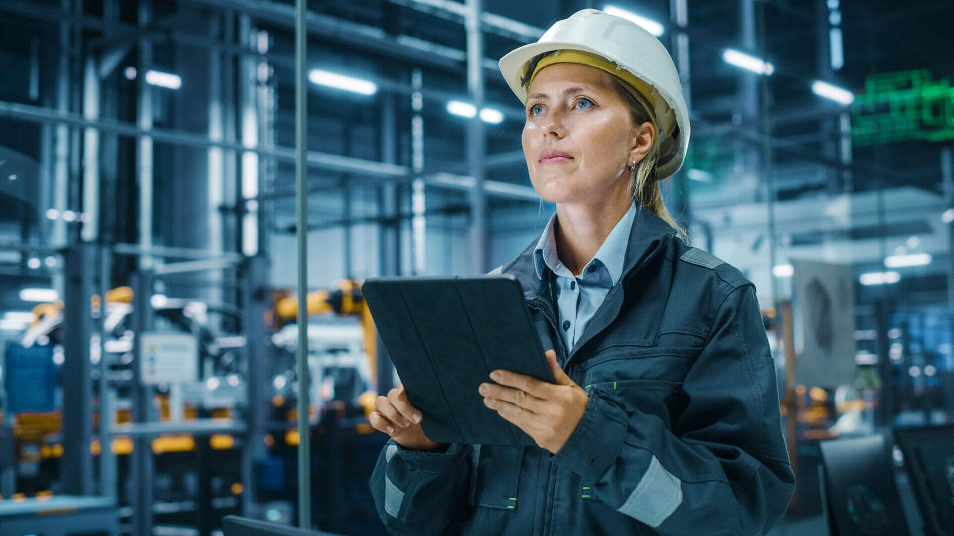 A woman in a hard hat is holding a tablet in a factory.